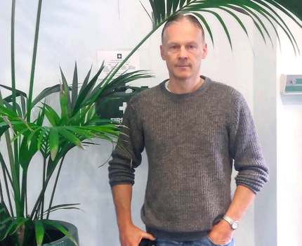 Man in gray sweater standing next to potted plant. Scott is a volunteer at Quotient Sciences.