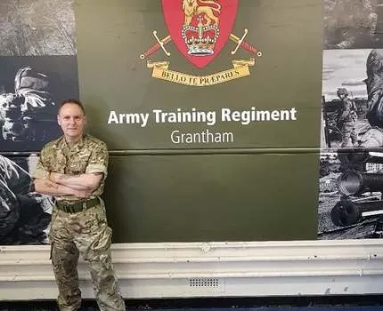 Photo of a man in an army uniform with green and tan colours camouflage print standing in front of a green sign that says Army Training Regiment